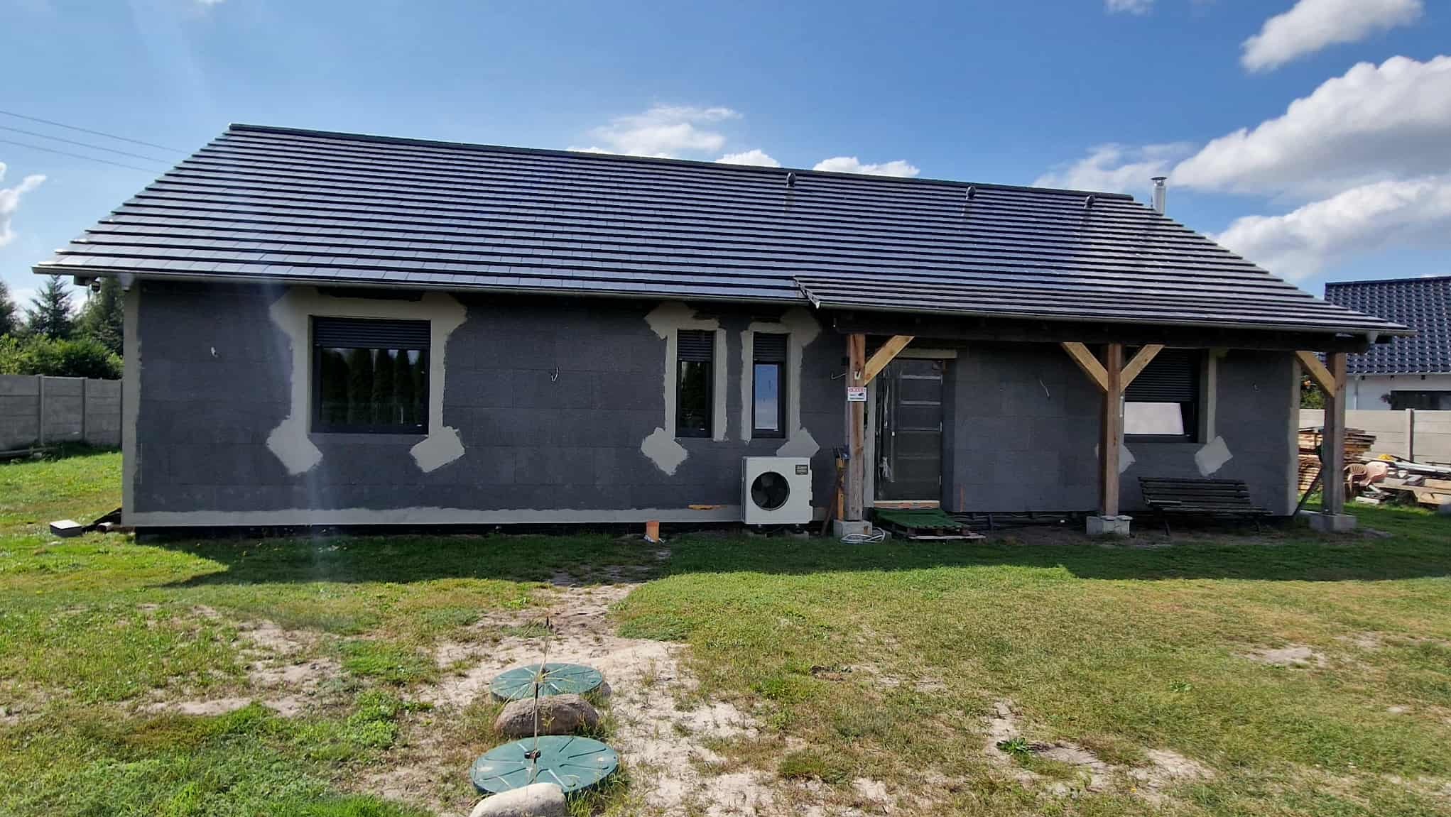 Dom w trakcie ocieplania: szara elewacja z widocznymi łatami kleju wokół okien, czarny dach, klimatyzator na zewnątrz. Widoczne elementy ogrodu i nieba.