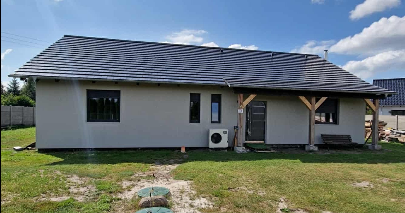 Elewacja domu jednorodzinnego z szarą fasadą, ciemną dachówką i drewnianą wiatą. Widoczny klimatyzator na ścianie i zadbany trawnik.