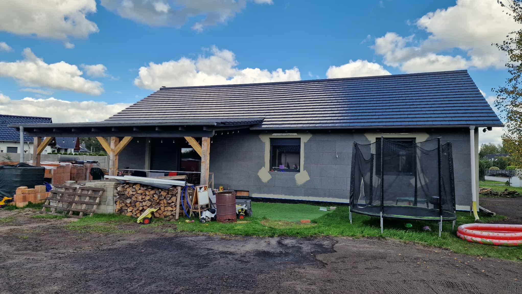 Dom w trakcie ocieplania ścian zewnętrznych, widoczne szare bloczki i fragmenty ocieplenia wokół okna. Nowy dach z ciemnej dachówki. Na pierwszym planie zabawki.