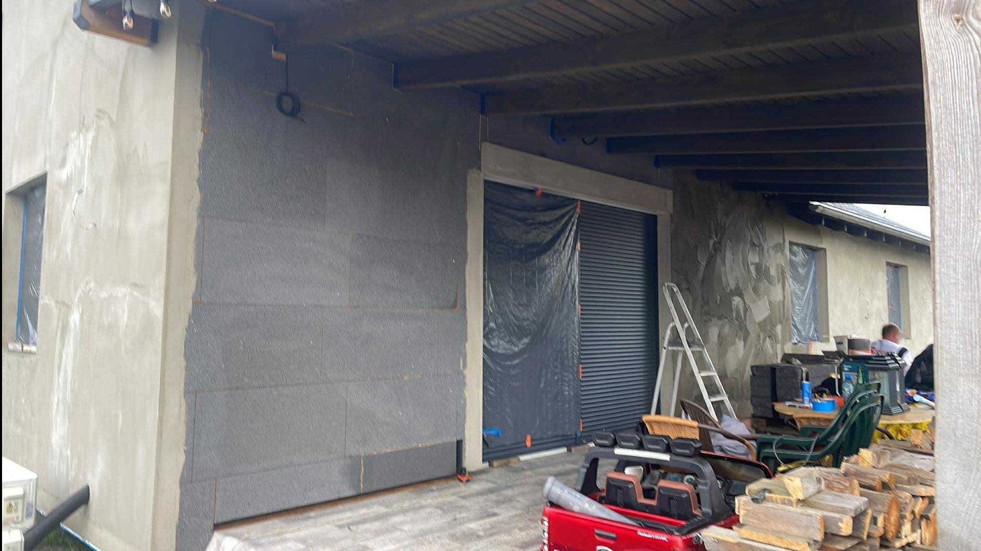 Ocieplanie ściany zewnętrznej budynku z widoczną warstwą izolacyjną i fragmentem elewacji po nałożeniu tynku. Częściowo zasłonięta brama garażowa.