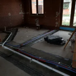 Wnętrze w surowym stanie z widocznymi, nowo ułożonymi rurami instalacji wodnej i grzewczej w kolorach białym, czerwonym i niebieskim, biegnącymi wzdłuż ścian na przygotowanej podłodze.