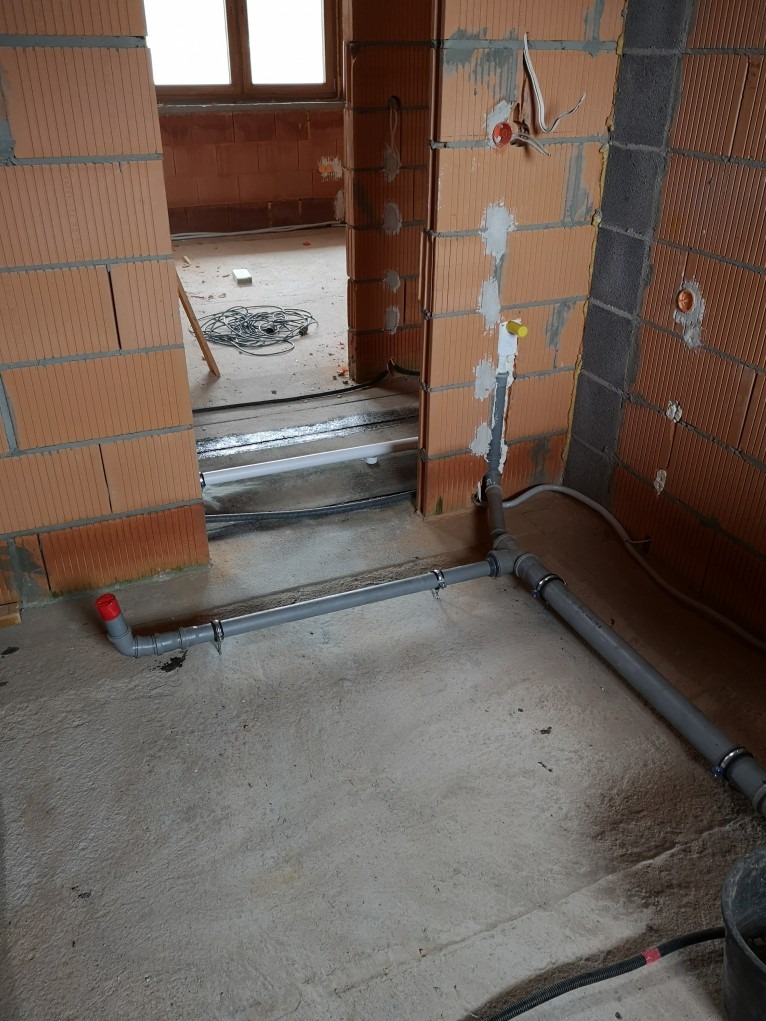 Szare rury kanalizacyjne z obejmami mocującymi ułożone na surowym betonowym podłożu w budynku w trakcie budowy, widoczne nieotynkowane ściany z czerwonej cegły i otwory na instalacje elektryczne.