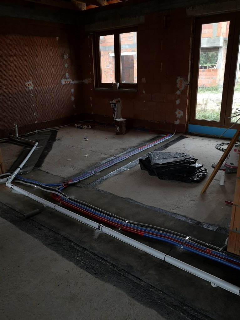Wnętrze w surowym stanie z widocznymi, nowo ułożonymi rurami instalacji wodnej i grzewczej w kolorach białym, czerwonym i niebieskim, biegnącymi wzdłuż ścian na przygotowanej podłodze.