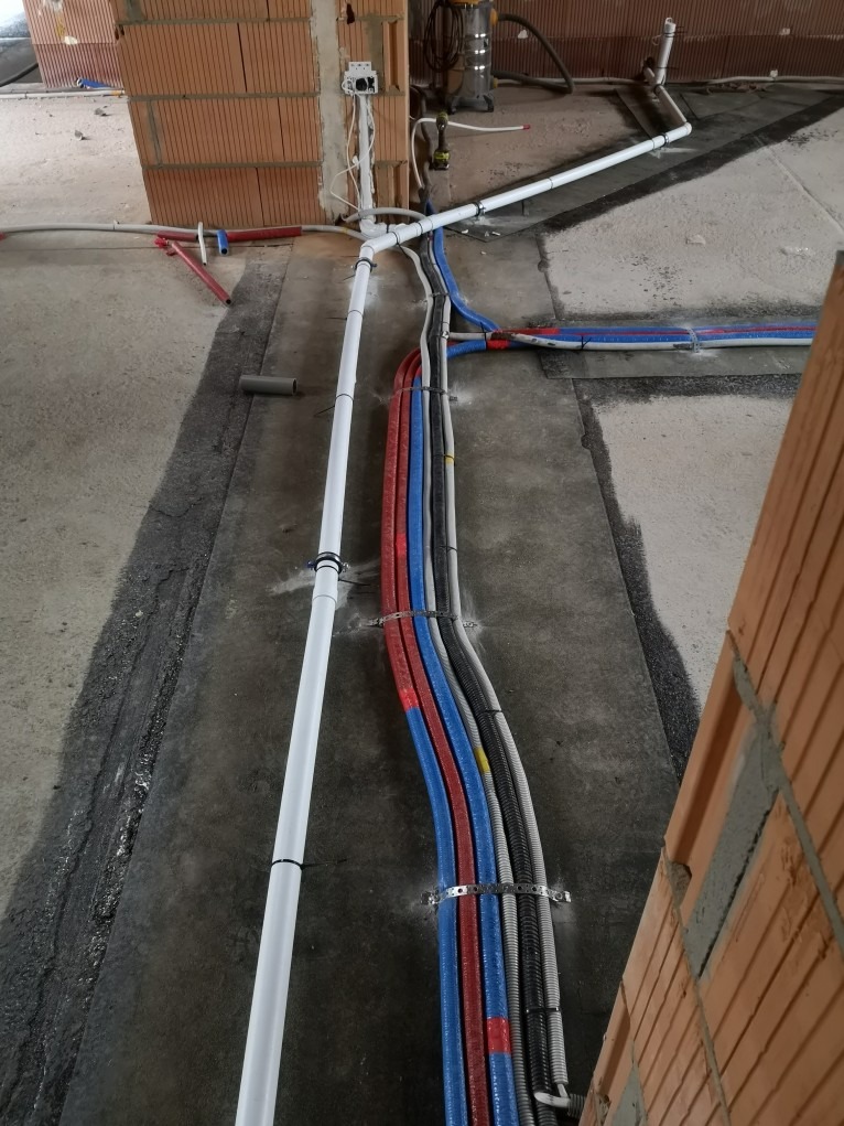Instalacja rur wodnych i kanalizacyjnych w surowym stanie, rury prowadzone po podłodze, mocowane metalowymi obejmami, widoczne przewody elektryczne i fragment ściany z cegły.