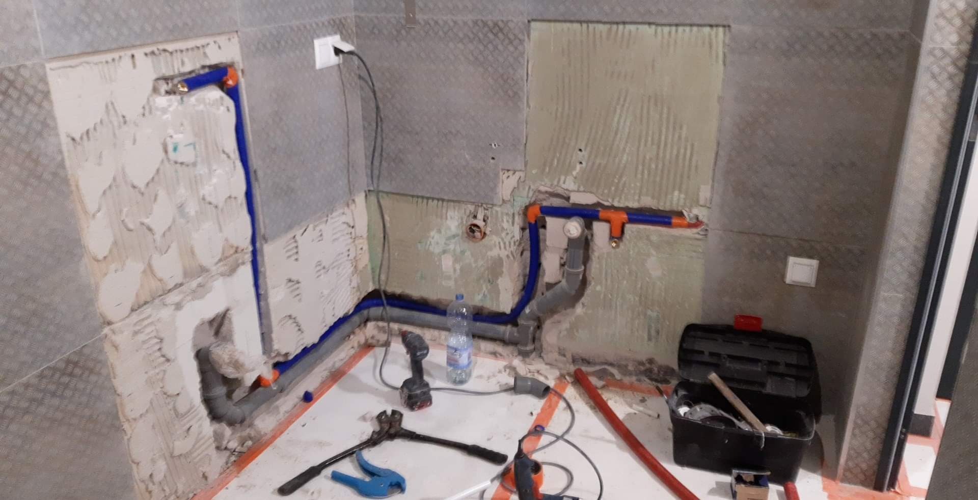 Niedokończona łazienka w trakcie prac hydraulicznych: widoczne rury, narzędzia, częściowo położone płytki i odsłonięte ściany.