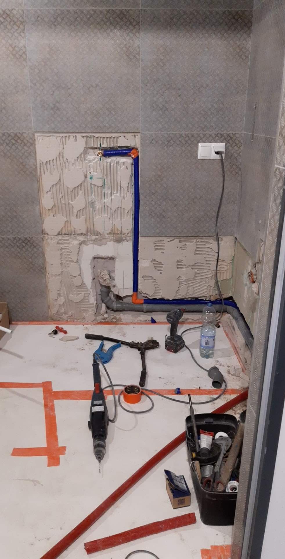 Remont łazienki w trakcie: widoczne fragmenty instalacji hydraulicznej z rur PCV w kolorze niebieskim i szarym, narzędzia hydrauliczne, wiertarka akumulatorowa, butelka wody, ślady po demontażu...