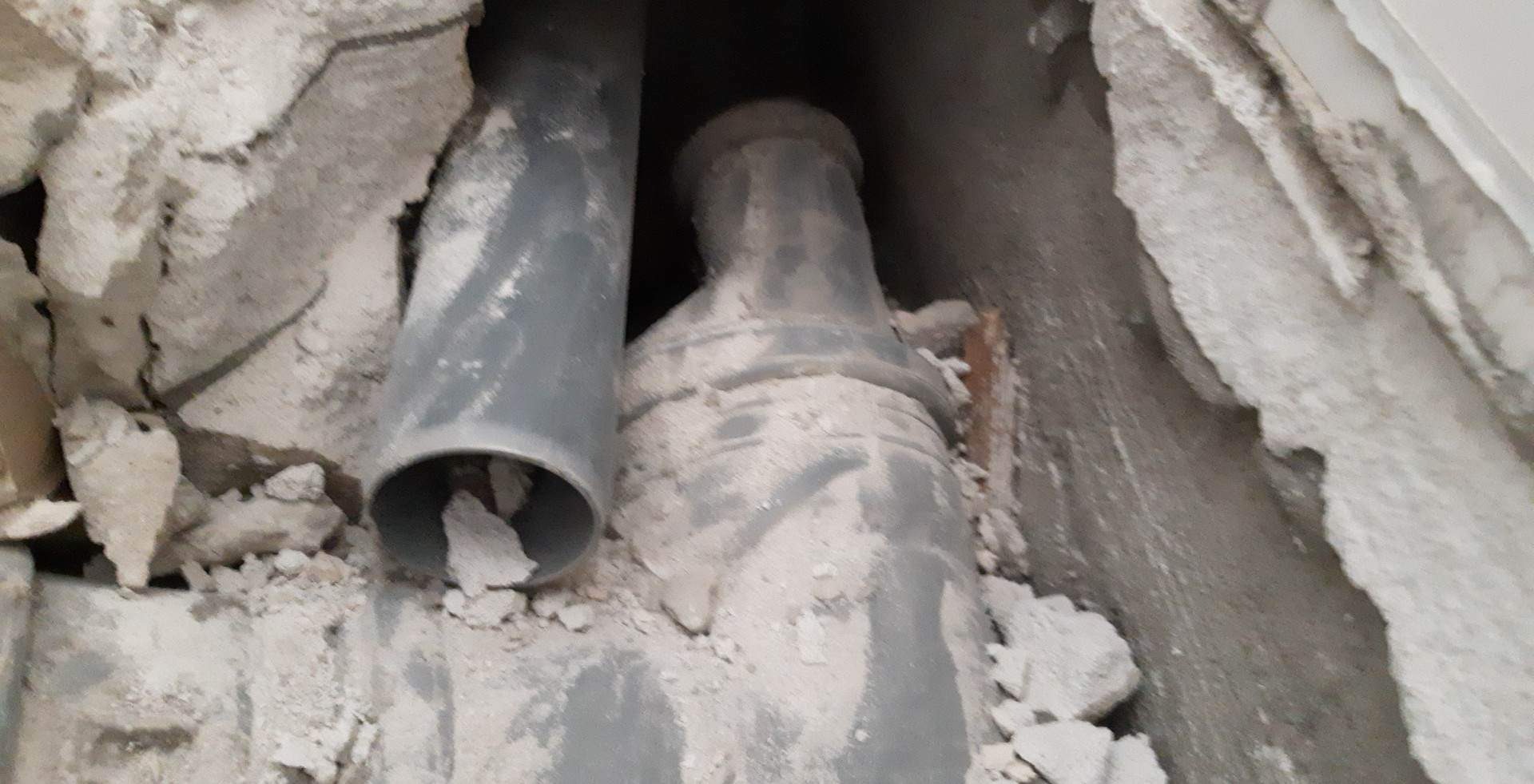 Szare rury kanalizacyjne w trakcie montażu w ścianie z gruzem i resztkami zaprawy, widoczne połączenia i fragment ściany.