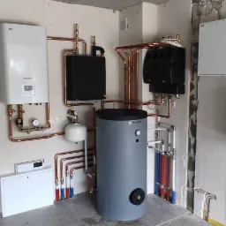 Kompleksowa instalacja grzewcza w surowym wnętrzu: biały piec gazowy LG, czarny wymiennik ciepła, szary zbiornik Trinity, miedziane rury i dwie białe skrzynki elektryczne na ścianie.