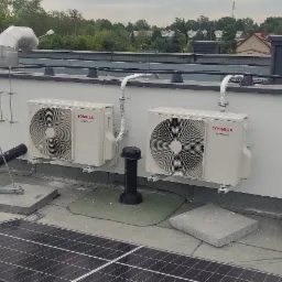 Dwa klimatyzatory Toshiba Inverter zamontowane na dachu budynku obok paneli słonecznych, przewody w izolacji, widok na zieleń w tle.