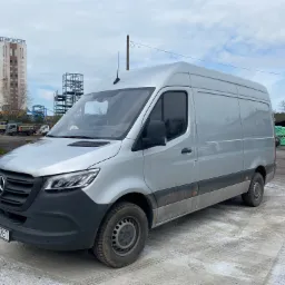 Srebrny samoch&oacute;d dostawczy Mercedes-Benz Sprinter na placu, w tle budynek z napisem 'IIWOMENI' i składowane worki z zieloną etykietą.