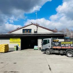 Budynek biura sprzedaży z zapasem worków i samochodem dostawczym z reklamą na pace, widoczne panele słoneczne na dachu.