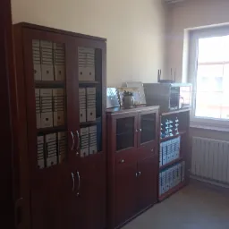 Biuro z ciemnymi drewnianymi regałami wypełnionymi segregatorami z dokumentami, na środkowym regale ramki ze zdjęciami i roślina doniczkowa, w tle okno.