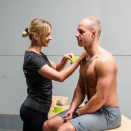 Kobieta aplikuje zieloną taśmę kinesiotaping na ramię mężczyzny siedzącego na leżance.