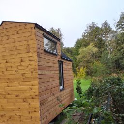 SMAL HOUSE - Zewnętrzny widok małego, drewnianego domu z dwoma oknami, otoczonego zielenią i drzewami.