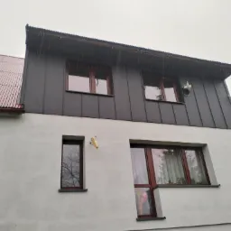 Dwukondygnacyjny budynek z szarą, metalową elewacją na górnej kondygnacji i białym tynkiem na dole, okna z brązowymi ramami, widoczny fragment dachu z blachy trapezowej i antena satelitarna...