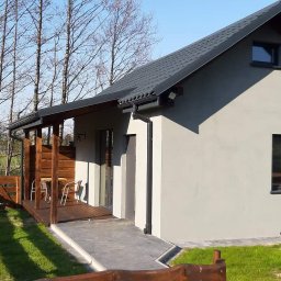 SMAL HOUSE - Gotowy dom szkieletowy z drewnianym tarasem i ogrodzeniem na tle drzew bez liści i zielonej trawy.