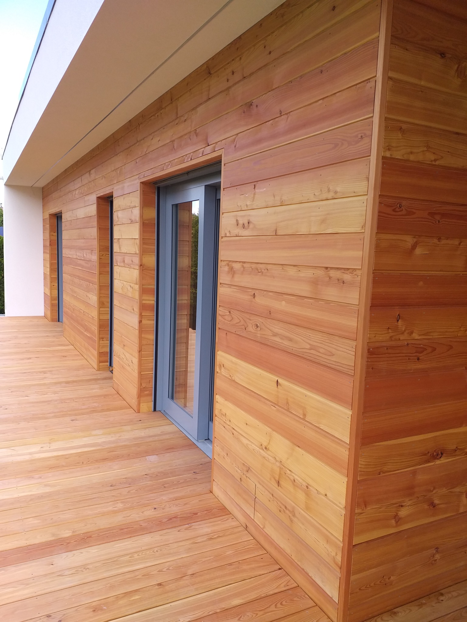 Elewacja budynku wykończona drewnianymi panelami, z widoczną drewnianą podłogą tarasu i szarymi ramami okiennymi.