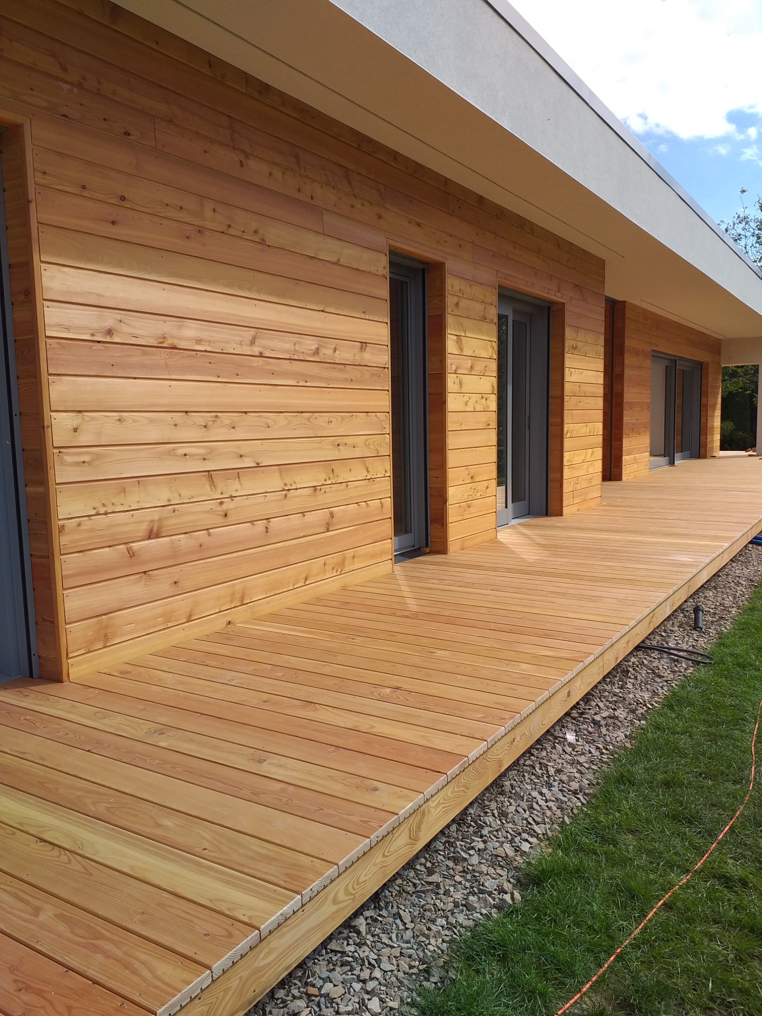 Elewacja domu z drewnianą okładziną i tarasem z desek kompozytowych, widoczne szare ramy okienne i minimalistyczny design budynku.