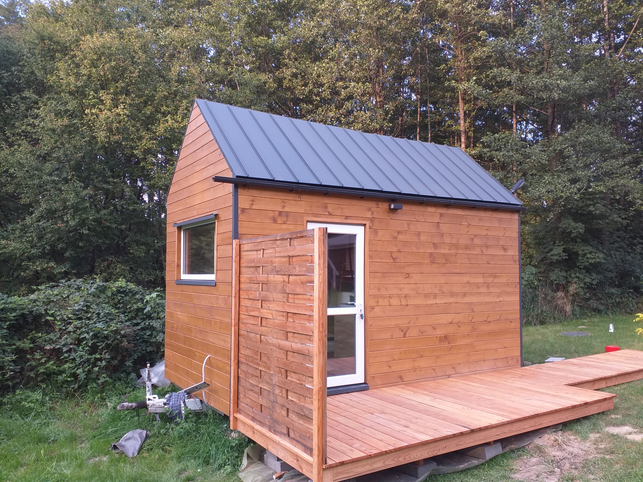 Mały drewniany domek z szarą blachą na dachu i drewnianym tarasem, widok z zewnątrz, w tle zieleń drzew.
