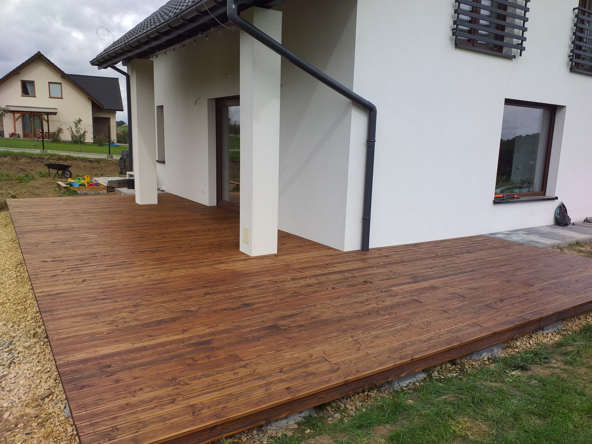 Drewniany taras przylegający do białego domu z ciemną rynną, widok na drugi dom w tle.