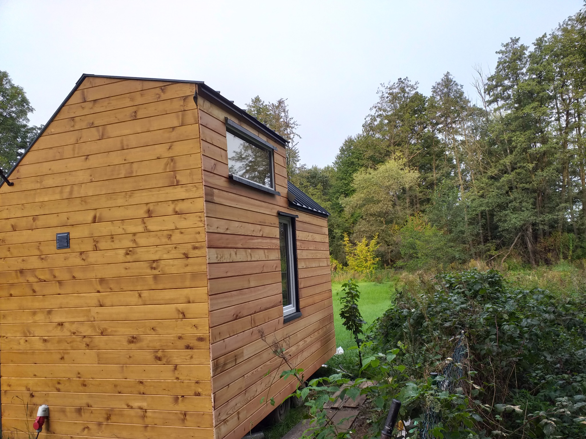 Zewnętrzny widok małego, drewnianego domu z dwoma oknami, otoczonego zielenią i drzewami.