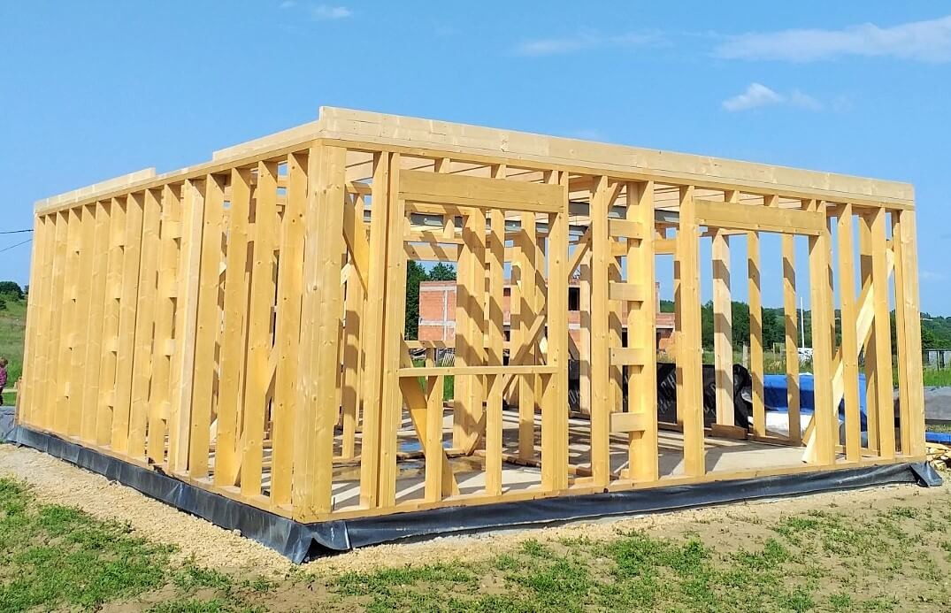 Surowa drewniana konstrukcja budynku z widocznymi słupkami i belkami, osadzona na fundamencie z czarną izolacją, na tle błękitnego nieba i zieleni.