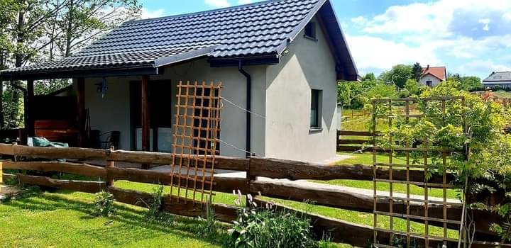 Niewielki, parterowy dom z szarą elewacją i ciemną dachówką, ogrodzony niskim, drewnianym płotem na tle zieleni.