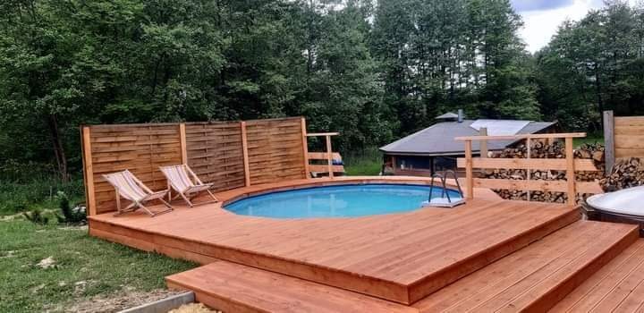 Owalny basen z błękitną wodą otoczony drewnianym tarasem i płotem z desek na tle zieleni drzew i małego domku.