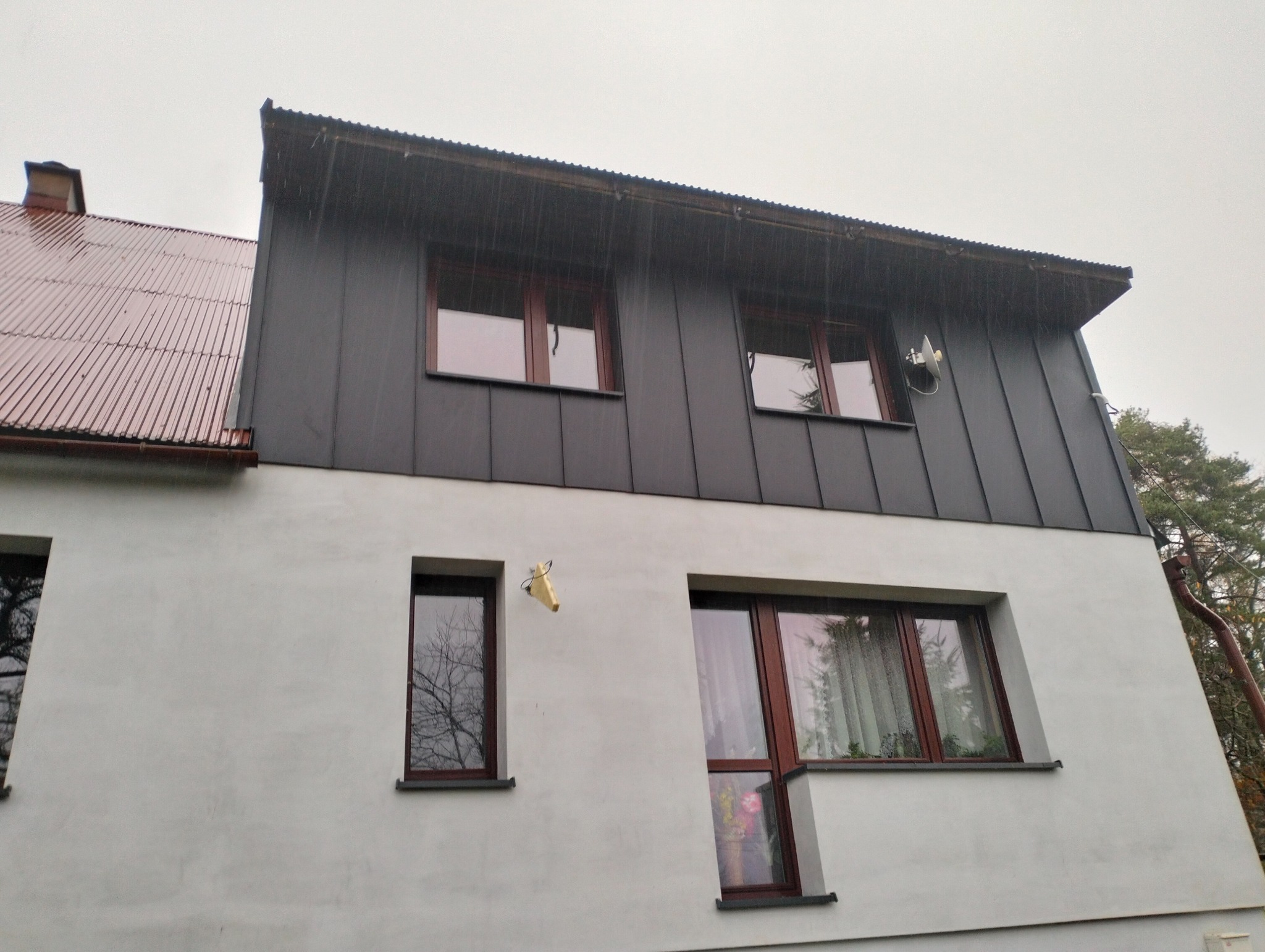 Dwukondygnacyjny budynek z szarą, metalową elewacją na górnej kondygnacji i białym tynkiem na dole, okna z brązowymi ramami, widoczny fragment dachu z blachy trapezowej i antena satelitarna...