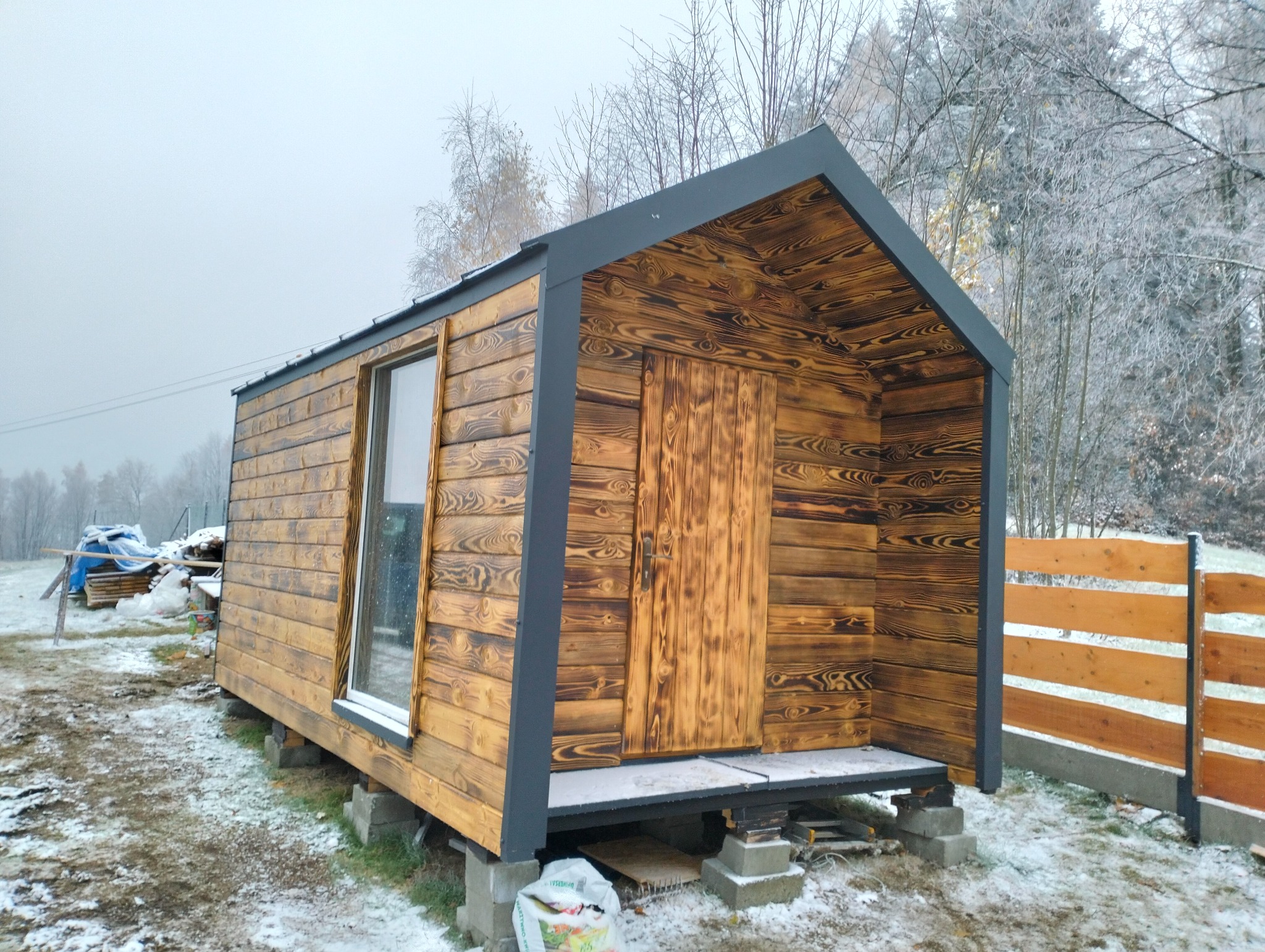 Nowoczesny dom szkieletowy z palonego drewna, z czarnymi elementami wykończeniowymi, posadowiony na betonowych bloczkach, zimowy krajobraz w tle.