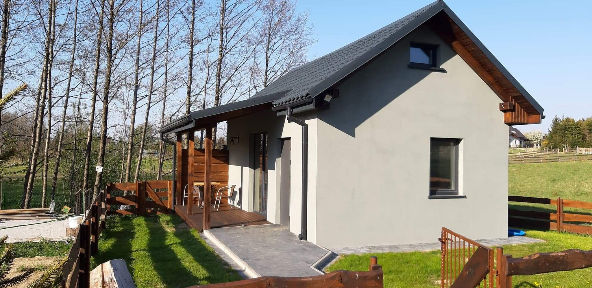 Gotowy dom szkieletowy z drewnianym tarasem i ogrodzeniem na tle drzew bez liści i zielonej trawy.