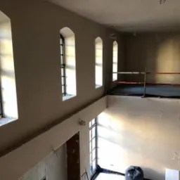 Wnętrze dwupoziomowego pomieszczenia w trakcie prac wykończeniowych ścian, widoczne trzy okna z łukowym wykończeniem, zabezpieczone podłogi na antresoli i worki z odpadami budowlanymi.