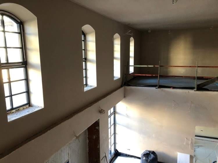 Wnętrze dwupoziomowego pomieszczenia w trakcie prac wykończeniowych ścian, widoczne trzy okna z łukowym wykończeniem, zabezpieczone podłogi na antresoli i worki z odpadami budowlanymi.