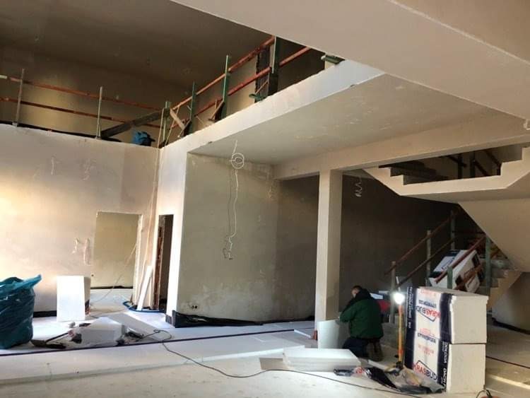 Wnętrze w trakcie prac wykończeniowych: ściany po wstępnym nałożeniu warstwy, widoczne rusztowania, materiały izolacyjne i pracownik w zielonej kurtce.