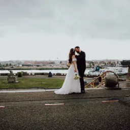 Para młoda w objęciach na tle panoramy miasta z diabelskim młynem, panna młoda trzyma bukiet białych r&oacute;ż.
