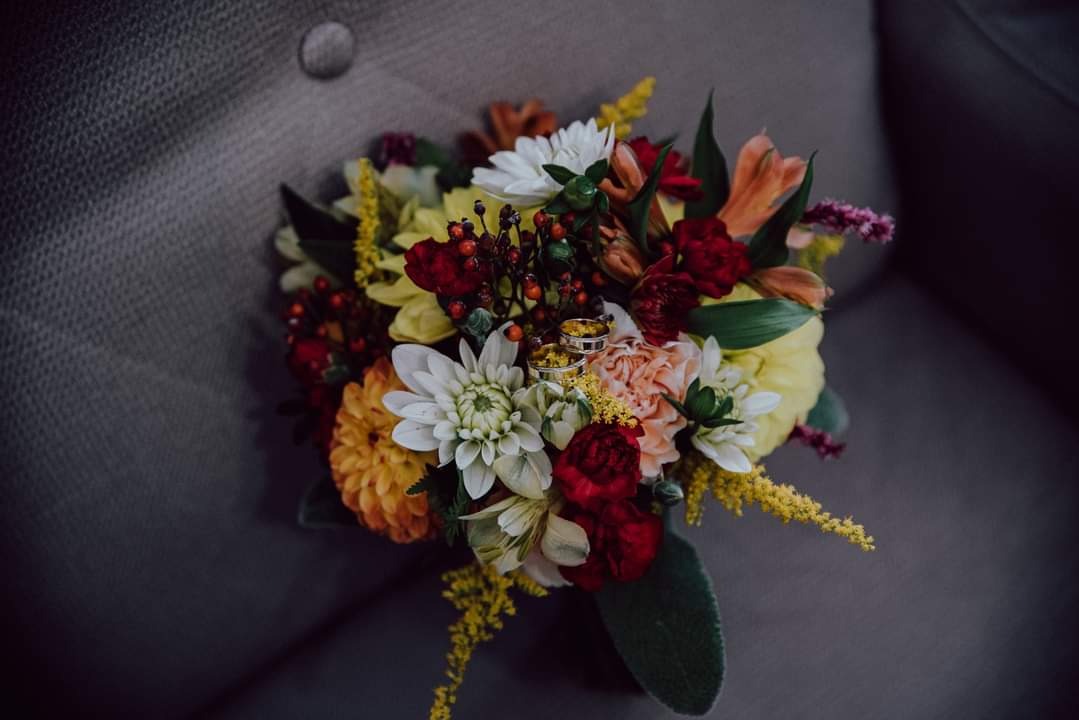 Złote obrączki ślubne spoczywają na bukiecie z białych, żółtych, czerwonych i pomarańczowych kwiatów, ułożonym na szarym, tapicerowanym fotelu.
