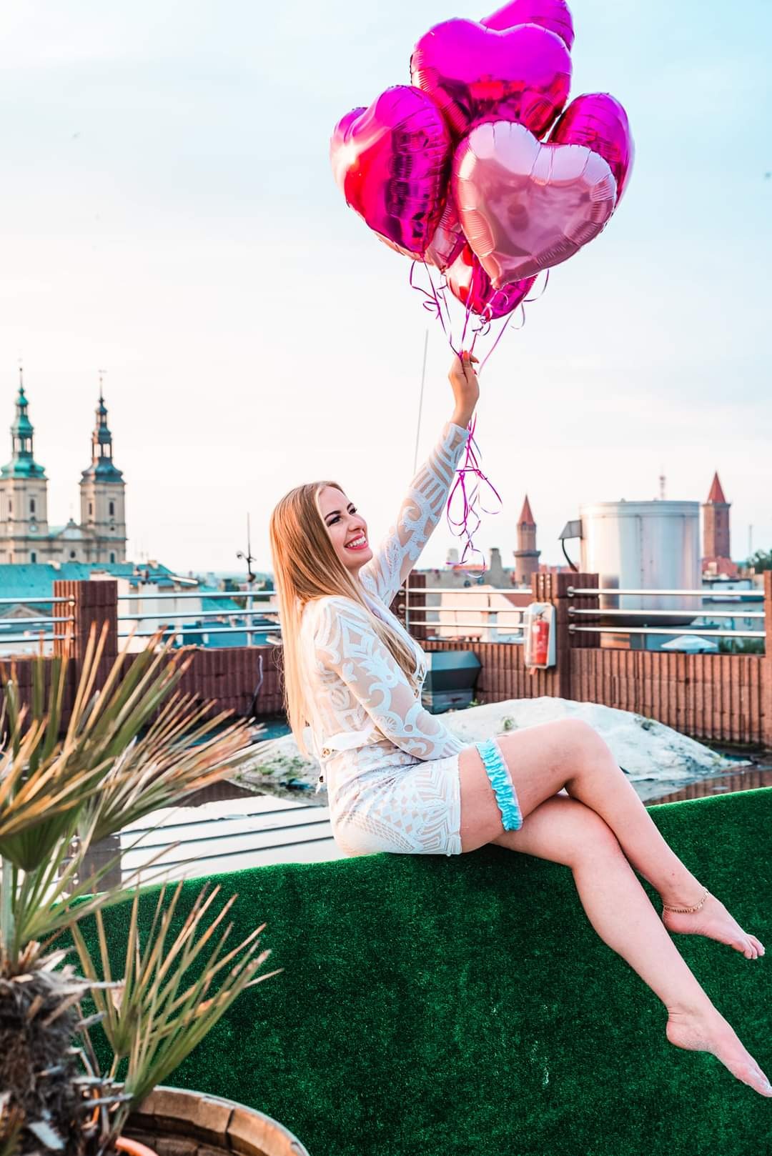 Uśmiechnięta blondynka w koronkowej sukience i niebieskiej podwiązce siedzi na zielonym dachu z widokiem na miasto i trzyma bukiet różowych balonów w kształcie serc.