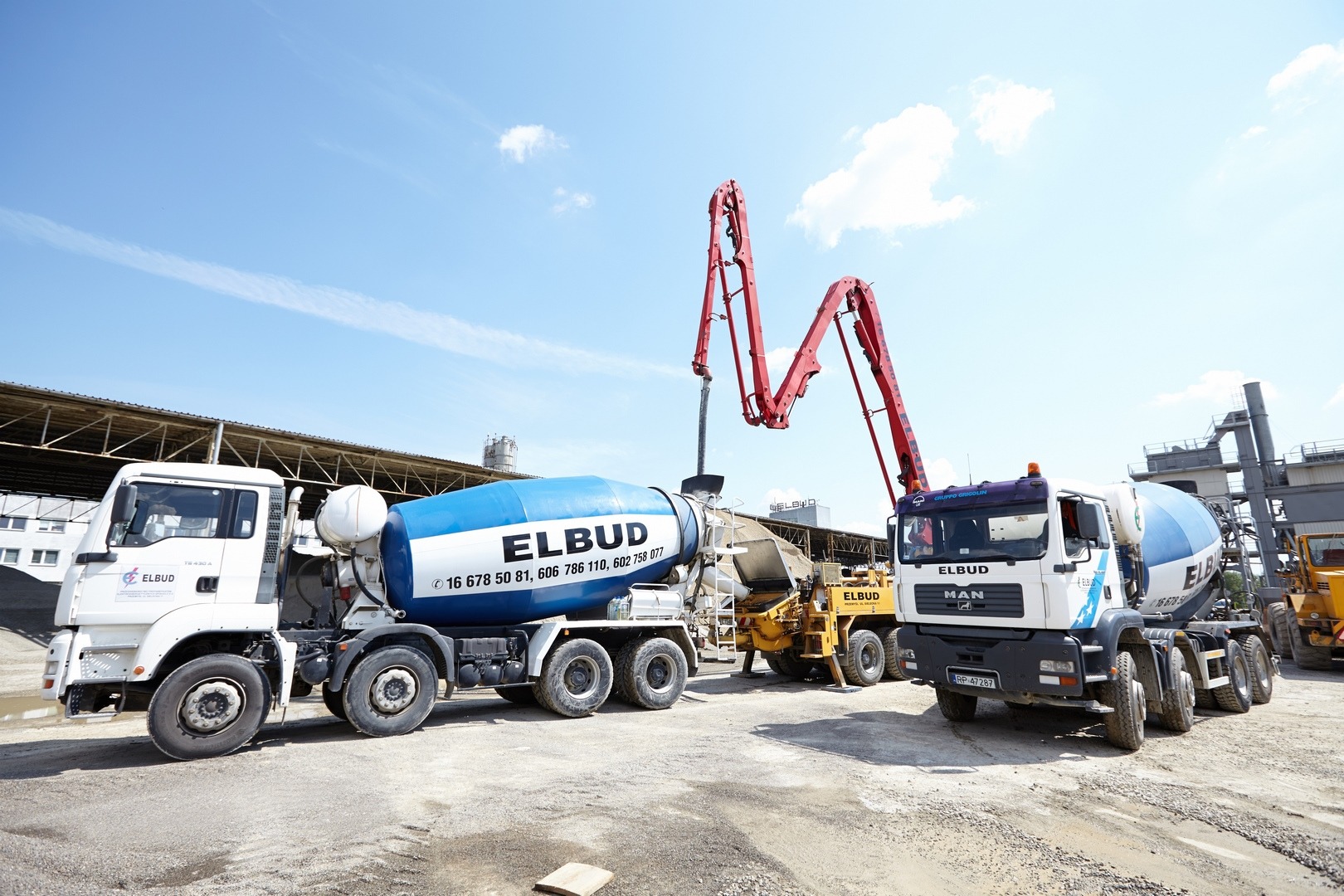Dwa białe betonomieszarki firmy Elbud z niebieskimi bębnami oraz czerwona pompa do betonu na placu budowy w słoneczny dzień.