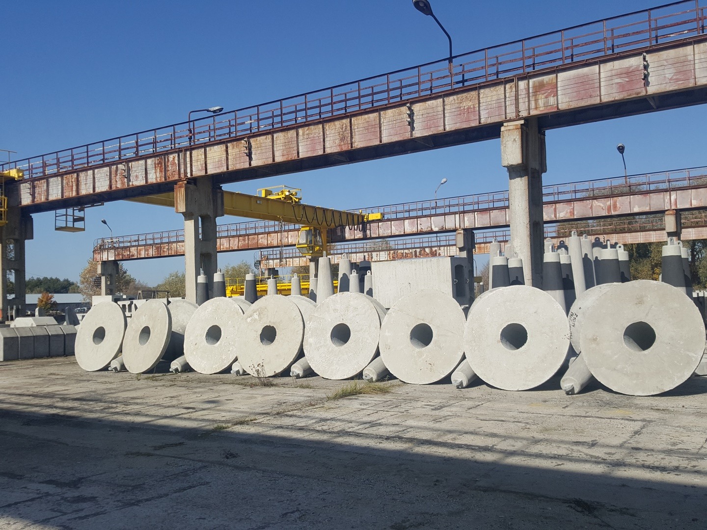 Rząd betonowych kręgów z otworem, ułożonych na placu pod suwnicą, w tle inne elementy betonowe i błękitne niebo.