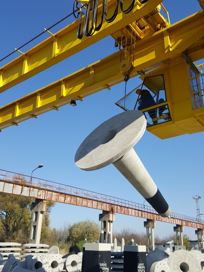 Żółty dźwig przenosi betonowy element w kształcie grzyba z czarnym oznaczeniem, na tle błękitnego nieba i konstrukcji nośnej.