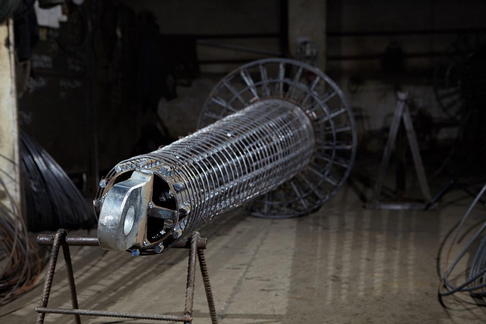 Metalowy szkielet cylindryczny z uchwytem, spawany z prętów, ustawiony na metalowym stojaku w warsztacie.