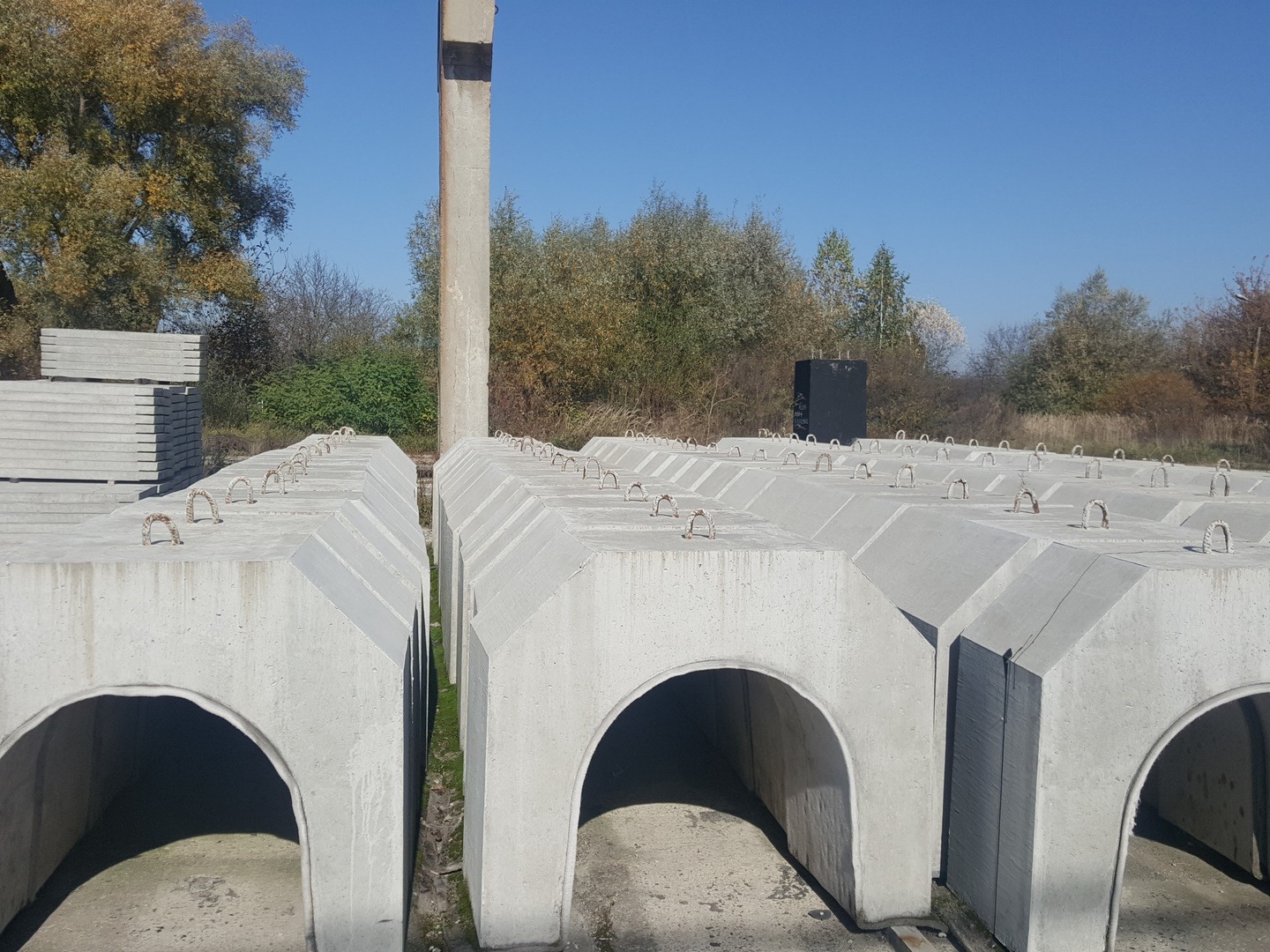 Szereg betonowych tuneli z metalowymi uchwytami na szczycie, składowanych na zewnątrz, z drzewami w tle i jasnym, błękitnym niebem.