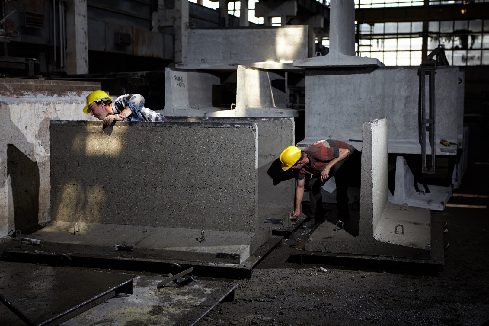 Dwóch pracowników w żółtych kaskach pracuje nad betonowymi prefabrykatami w hali produkcyjnej, jeden wygładza powierzchnię, drugi nadzoruje z góry.