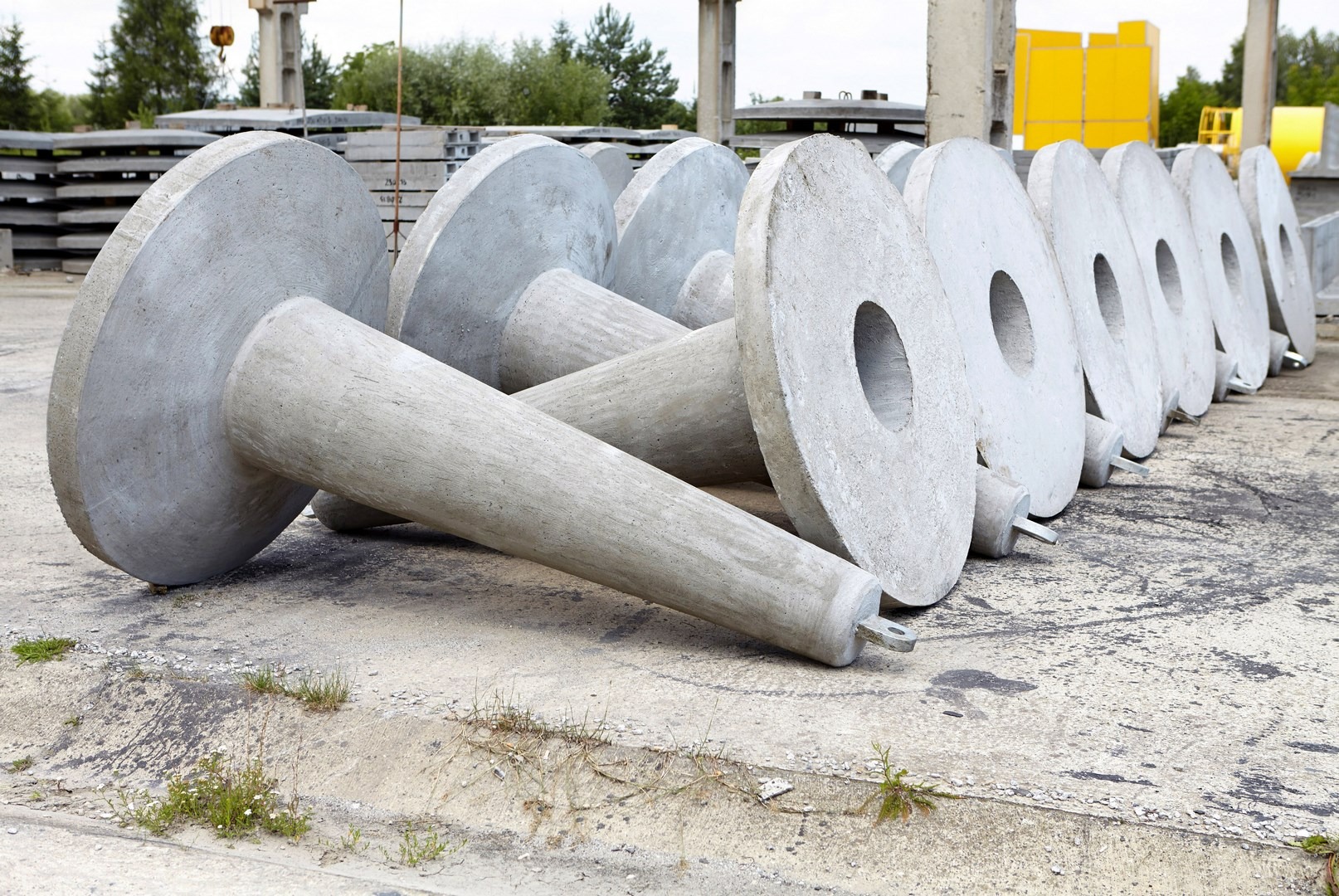 Szereg betonowych elementów o kształcie dysku z otworem i stożkową podstawą, leżących na betonowej powierzchni, z widocznymi śladami użytkowania i roślinnością w tle.