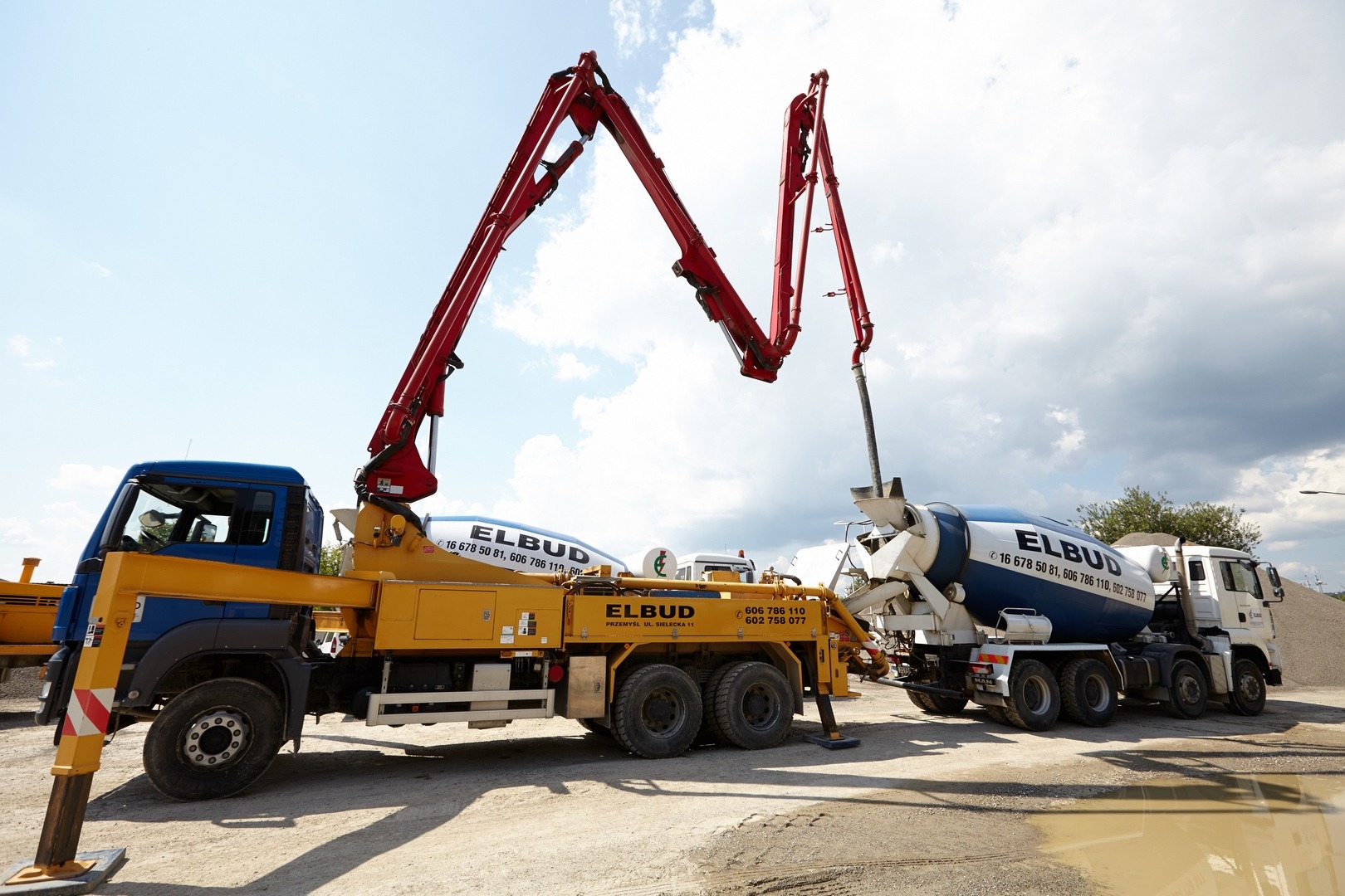 Żółto-niebieska betonomieszarka z wysięgnikiem pompy do betonu na placu budowy, obok stoi biała betonomieszarka, obie z logo firmy Elbud.