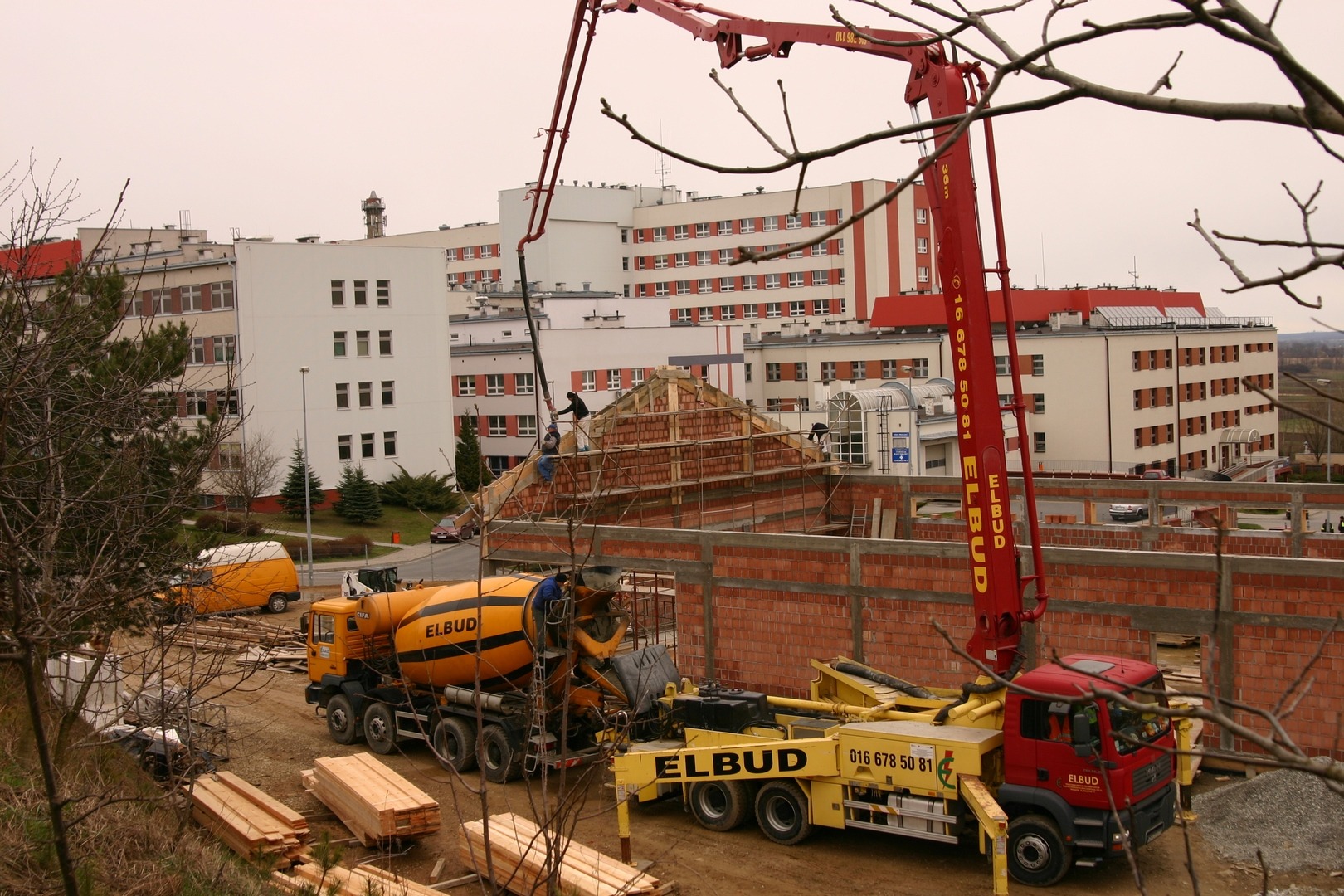 Żółta betonomieszarka i czerwony wysięgnik pompy do betonu na placu budowy, w tle budynek szpitala i robotnicy na rusztowaniu w trakcie murowania ściany.