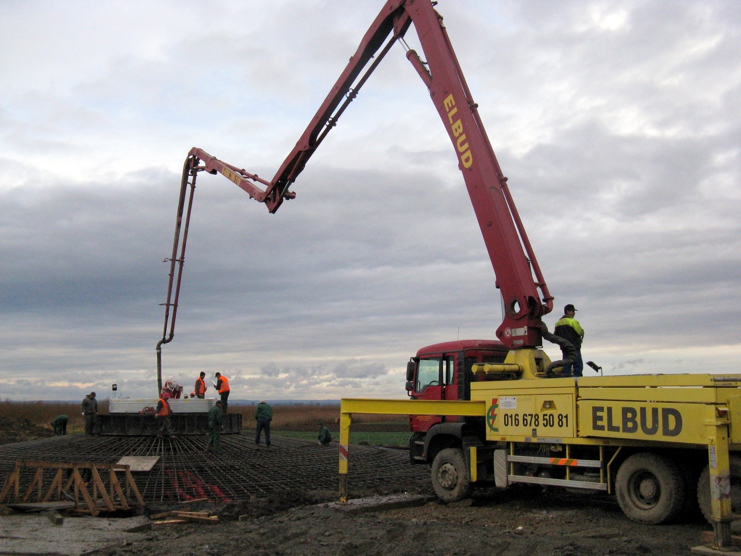 Żółto-czerwona pompa do betonu marki Elbud w trakcie wylewania betonu na zbrojenie fundamentu, widoczni pracownicy w odblaskowych kamizelkach, pochmurne niebo w tle.