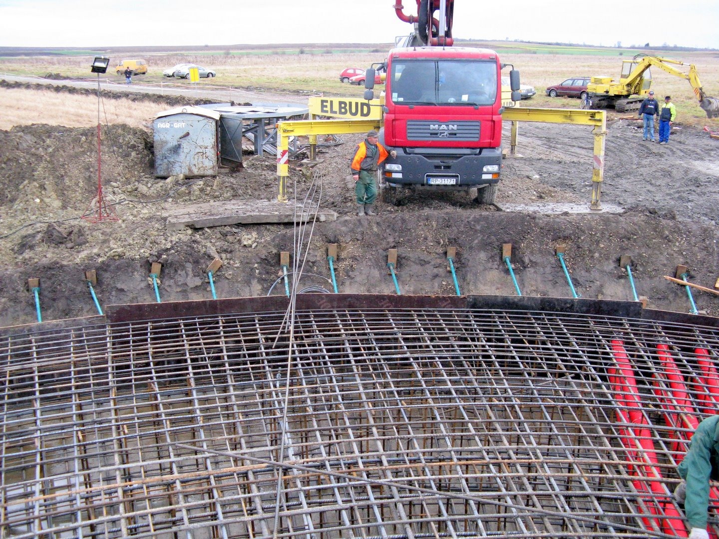 Wykonywanie zbrojenia pod fundament okrągłego zbiornika, z widocznym czerwonym betonopompą MAN, pracownikami w odblaskowych kamizelkach i koparką w tle, na placu budowy.