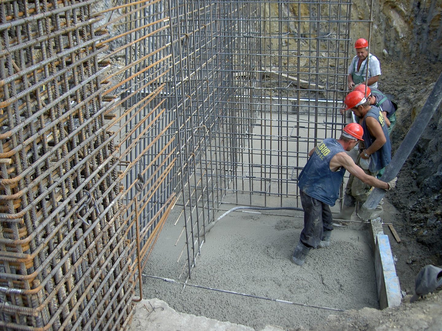 Wylewanie betonu do szalunku zbrojonego stalą, pracownicy w kaskach nadzorują proces, widoczna pompa do betonu i siatka zbrojeniowa.