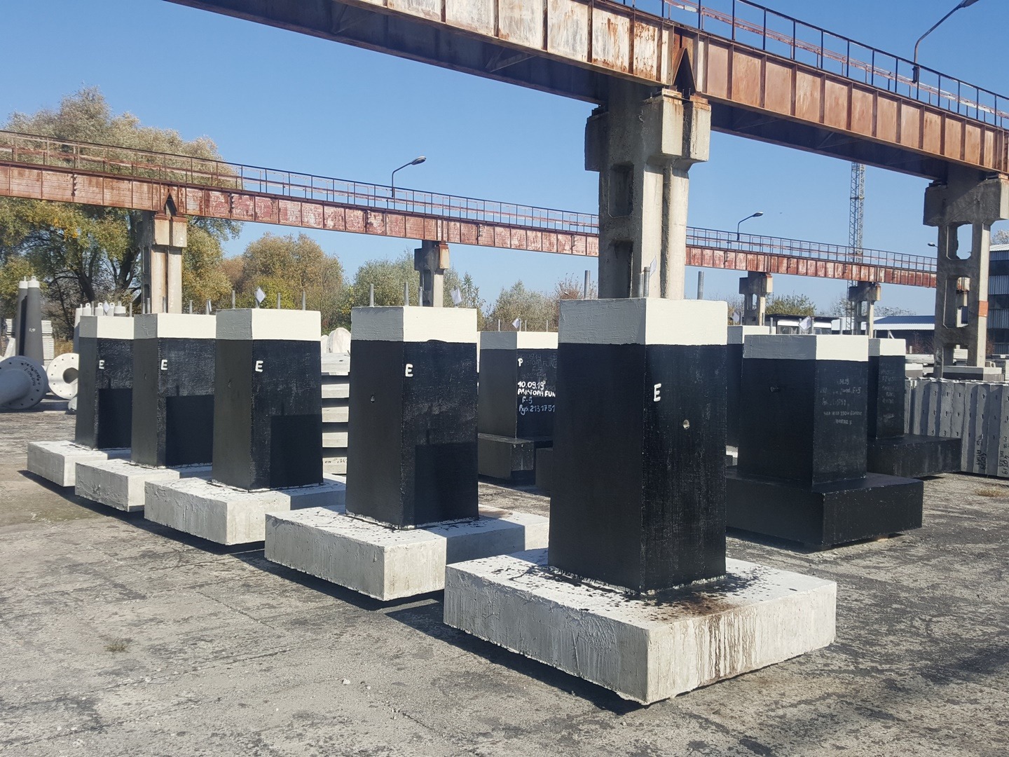 Rząd czarnych, betonowych prefabrykatów z białymi górnymi i dolnymi częściami, oznaczonych literą 'E', składowanych na placu budowy pod konstrukcją stalową suwnicy.